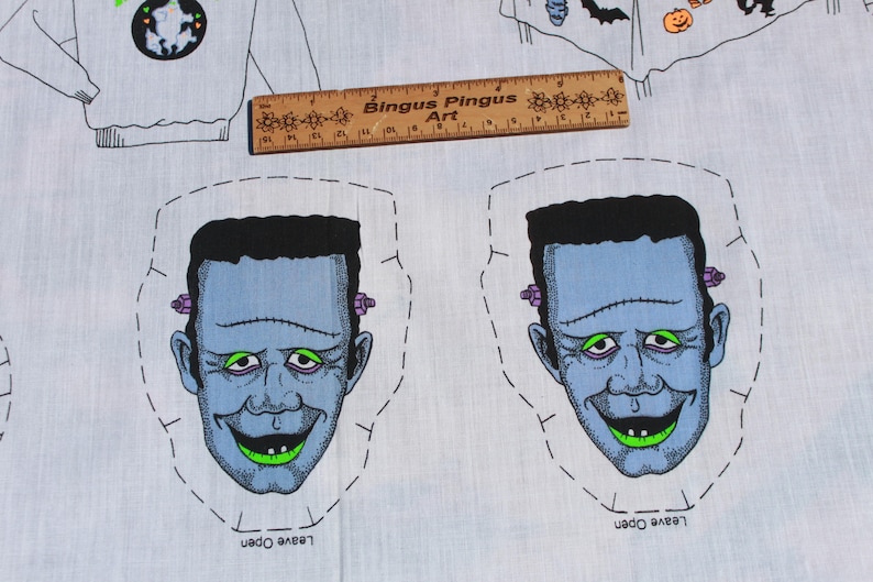 May include: A light blue fabric with two cartoon Frankenstein faces, each with black hair, green eyes, and a stitched mouth. The fabric also features a ruler with the text "Bingus Pingus Art" and Halloween-themed illustrations.