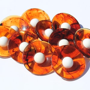 以下が含まれることがあります： 琥珀色の円形装飾品コレクション。中央に白い球体があります。オブジェクトは大理石模様で、白い表面に配置されています。全体的なテーマは装飾です。