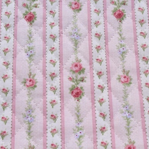 May include: Close-up of a quilted fabric with a floral pattern. The design features vertical stripes in pink and white, with rows of pink roses and small purple flowers. The fabric is stitched with a diamond pattern.