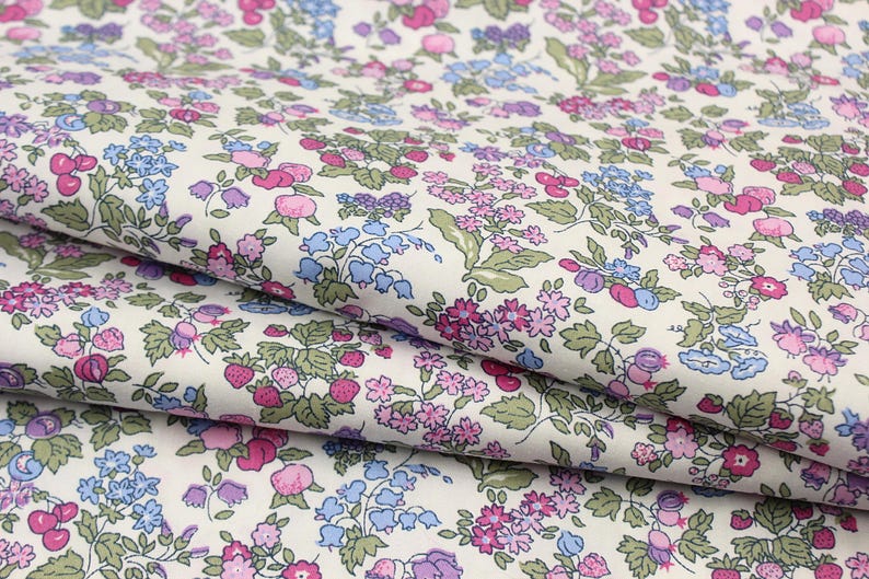 May include: A fabric featuring a repeating pattern of small pink, blue, and purple flowers interspersed with green leaves and red strawberries on an off-white background. The textile is folded to show the intricate botanical print and texture.