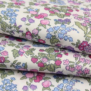 May include: A folded piece of fabric with an off-white background featuring a repeating pattern of small pink strawberries, blue bell-shaped flowers, and clusters of tiny pink and purple blossoms with green leaves arranged across the surface.