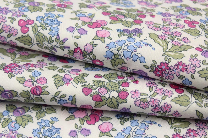 May include: A folded piece of fabric with an off-white background featuring a repeating pattern of small pink strawberries, blue bell-shaped flowers, and clusters of tiny pink and purple blossoms with green leaves arranged across the surface.