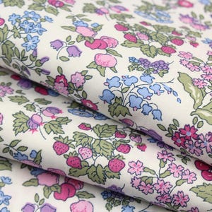 May include: A white fabric featuring a repeating botanical print of pink strawberries, purple berries, light blue bell-shaped flowers, and small pink blossoms with green leaves. The textile is folded in layers, showcasing the dense floral pattern.