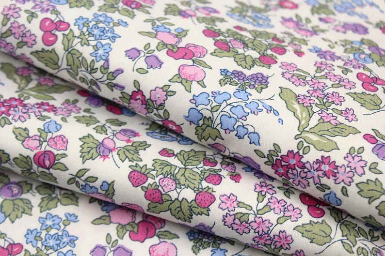 May include: A white fabric featuring a repeating botanical print of pink strawberries, purple berries, light blue bell-shaped flowers, and small pink blossoms with green leaves. The textile is folded in layers, showcasing the dense floral pattern.