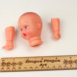 May include: A disassembled vintage doll with a peach-colored head, arms, and hands. The doll's head has blue eyes and red lips. A wooden ruler with the text "Bingus Pingus Art" is at the bottom.