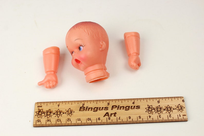 May include: A disassembled vintage doll with a peach-colored head, arms, and hands. The doll's head has blue eyes and red lips. A wooden ruler with the text "Bingus Pingus Art" is at the bottom.
