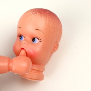 May include: A vintage plastic doll head and arm, featuring a peach-colored complexion, blue eyes, and rosy cheeks. The doll's arm is raised, with a finger touching the doll's mouth. The doll's hair is molded, and the head is cut at the neck.