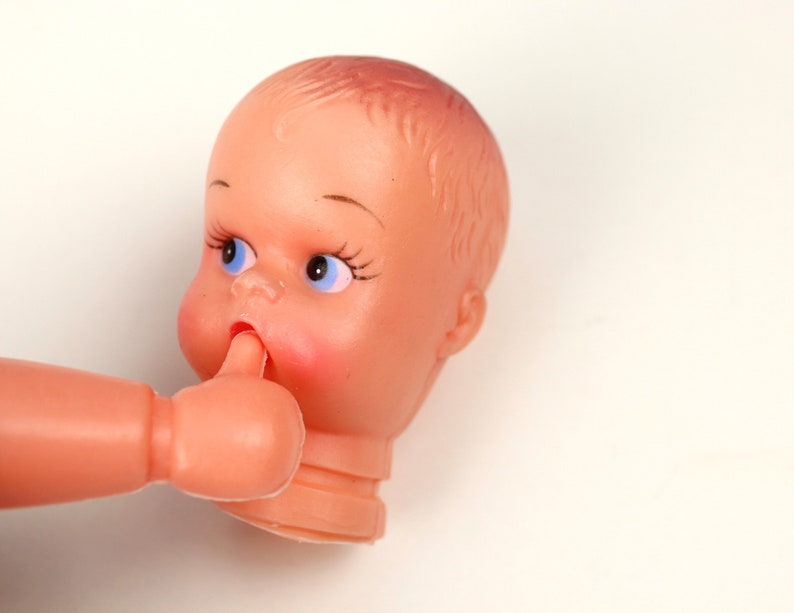 May include: A vintage plastic doll head and arm, featuring a peach-colored complexion, blue eyes, and rosy cheeks. The doll's arm is raised, with a finger touching the doll's mouth. The doll's hair is molded, and the head is cut at the neck.