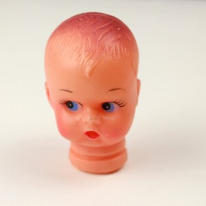 May include: A vintage doll head with a peach complexion, blue eyes, and a slightly open mouth. The doll head has painted eyelashes and eyebrows, with a hint of blush on the cheeks. The hair is a darker shade of pink.