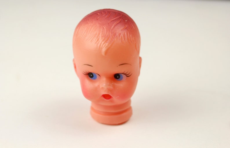 May include: A vintage doll head with a peach complexion, blue eyes, and a slightly open mouth. The doll head has painted eyelashes and eyebrows, with a hint of blush on the cheeks. The hair is a darker shade of pink.