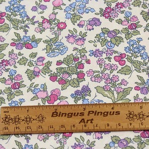 May include: A cream-colored fabric featuring a repeating botanical print of pink, purple, and blue flowers, green leaves, and red strawberries. A wooden ruler measuring 6 inches is placed horizontally across the bottom of the fabric pattern.