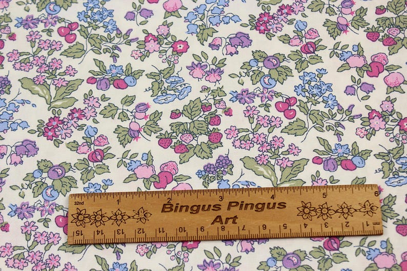 May include: A cream-colored fabric featuring a repeating botanical print of pink, purple, and blue flowers, green leaves, and red strawberries. A wooden ruler measuring 6 inches is placed horizontally across the bottom of the fabric pattern.