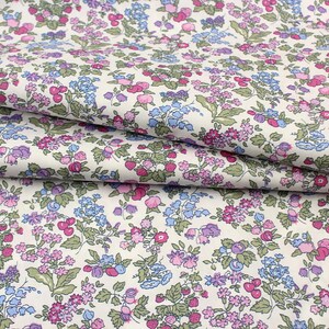 May include: A white fabric featuring a dense, repeating floral pattern with small pink, purple, and light blue flowers, along with green leaves and small red berries scattered throughout the design, shown in folded layers to display the print.