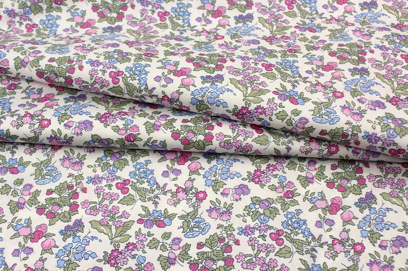 May include: A white fabric featuring a dense, repeating floral pattern with small pink, purple, and light blue flowers, along with green leaves and small red berries scattered throughout the design, shown in folded layers to display the print.