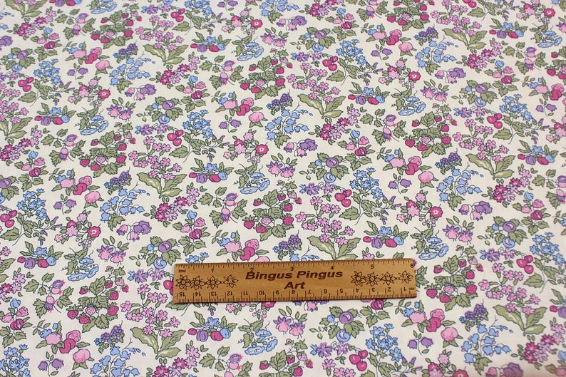 May include: A fabric with a repeating floral pattern featuring small pink, purple, and blue flowers with green leaves on a cream background. A wooden ruler labeled Bingus Pingus Art is placed across the fabric, showing a length of 6 inches.