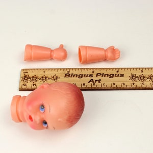 May include: A disassembled doll head and arms in a peach color, with blue eyes and a red mouth. A wooden ruler with the text "Bingus Pingus Art" is also present. The doll's head is approximately 2 inches tall.