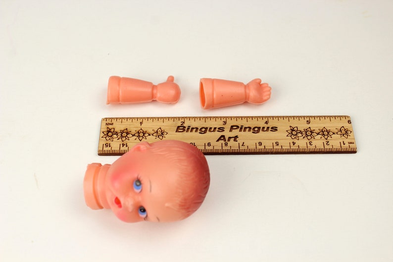 May include: A disassembled doll head and arms in a peach color, with blue eyes and a red mouth. A wooden ruler with the text "Bingus Pingus Art" is also present. The doll's head is approximately 2 inches tall.
