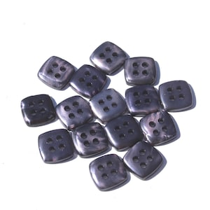May include: A collection of dark gray, square buttons with four holes each. The buttons have rounded corners and a slightly iridescent sheen, scattered on a white background. These buttons are suitable for sewing projects and garment construction.