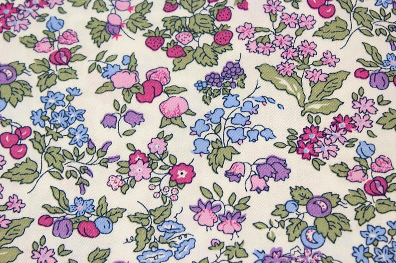 May include: A cream-colored fabric featuring a repeating botanical print of small strawberries, cherries, and various clusters of pink, purple, and light blue flowers with green leaves. The pattern is dense and evenly distributed across the surface.