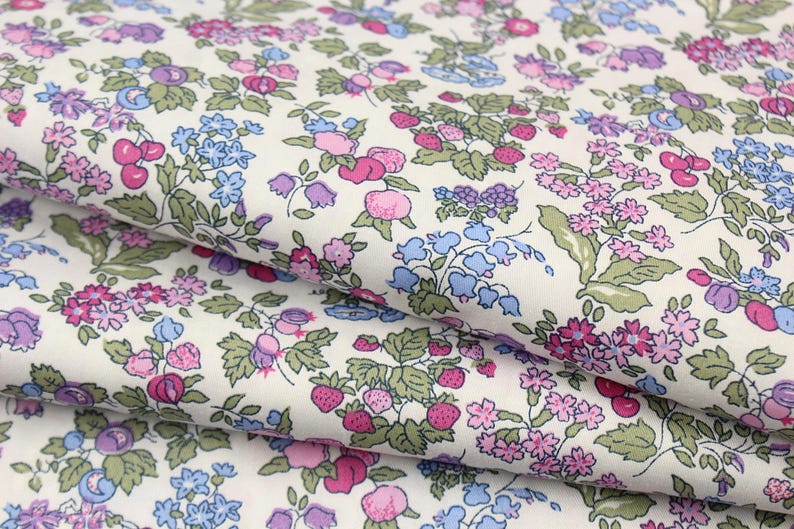 May include: A white fabric featuring a dense, repeating botanical print of small pink and purple flowers, light blue bell-shaped blossoms, green leaves, and clusters of red strawberries and cherries scattered across the surface in a continuous pattern.