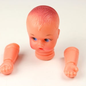 May include: A disassembled vintage doll head and arms. The doll head is peach-colored with blue eyes, painted eyelashes, and a red mouth. The arms are also peach-colored, with detailed hands. The doll's head and arms are separated.