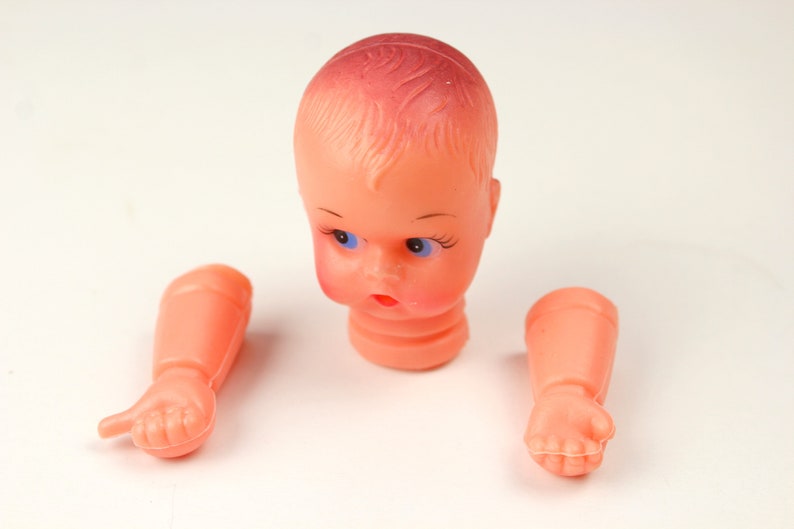 May include: A disassembled vintage doll head and arms. The doll head is peach-colored with blue eyes, painted eyelashes, and a red mouth. The arms are also peach-colored, with detailed hands. The doll's head and arms are separated.