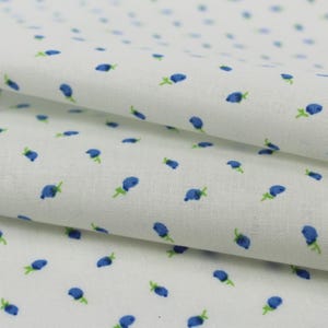 May include: White fabric with a repeating pattern of small blue floral designs with green stems. The fabric is folded, showing the texture and pattern in detail. The overall effect is delicate and suitable for various craft projects.