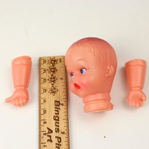 May include: A vintage doll head and two arms in a peach color, with a wooden ruler for scale. The doll head has blue eyes, red lips, and a slightly surprised expression. The ruler is marked in inches.