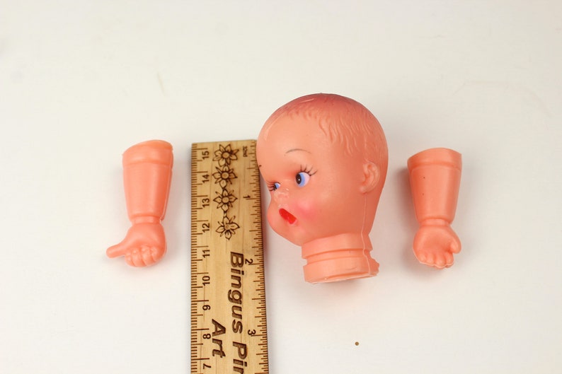 May include: A vintage doll head and two arms in a peach color, with a wooden ruler for scale. The doll head has blue eyes, red lips, and a slightly surprised expression. The ruler is marked in inches.