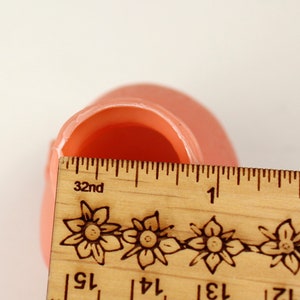 May include: A light peach-colored, rounded object with a hollow interior is measured with a wooden ruler. The ruler, marked in inches, shows the object's width. The ruler has floral designs and the number 32nd printed on it.