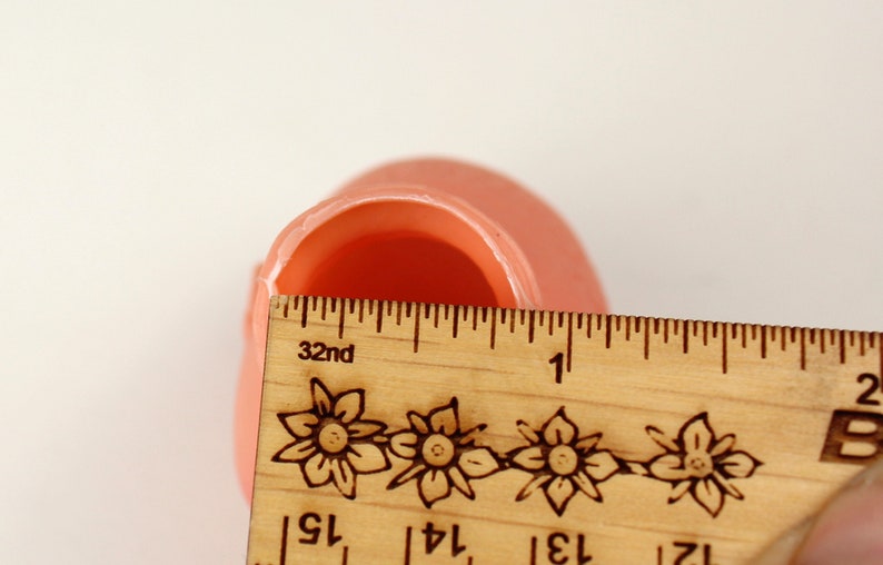 May include: A light peach-colored, rounded object with a hollow interior is measured with a wooden ruler. The ruler, marked in inches, shows the object's width. The ruler has floral designs and the number 32nd printed on it.