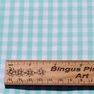 May include: A close-up of a white and light blue gingham fabric with a ruler measuring 4 inches. The ruler has the words "Bingus Pingus Art" printed on it.