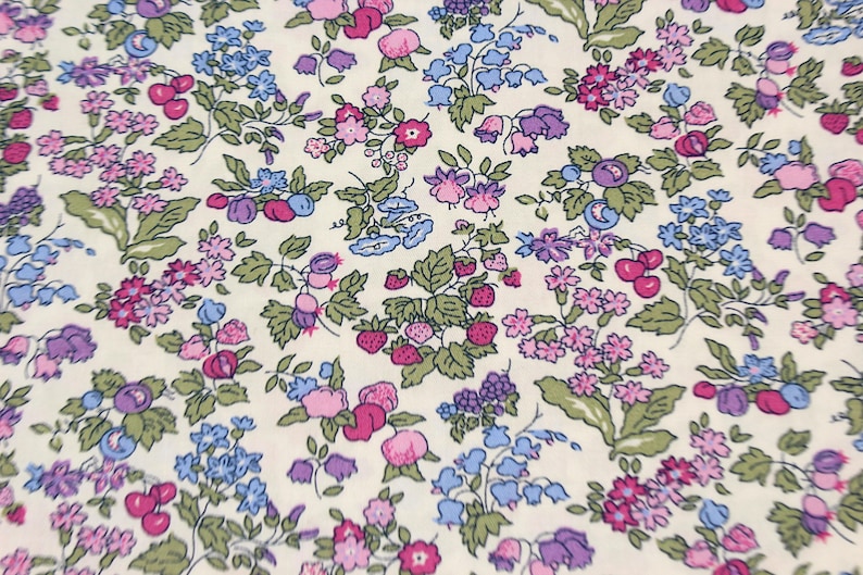 May include: A cream-colored fabric featuring a dense, repeating botanical print. The pattern consists of small pink, purple, and blue flowers, green leaves, and clusters of red strawberries and dark blue berries scattered across the surface.