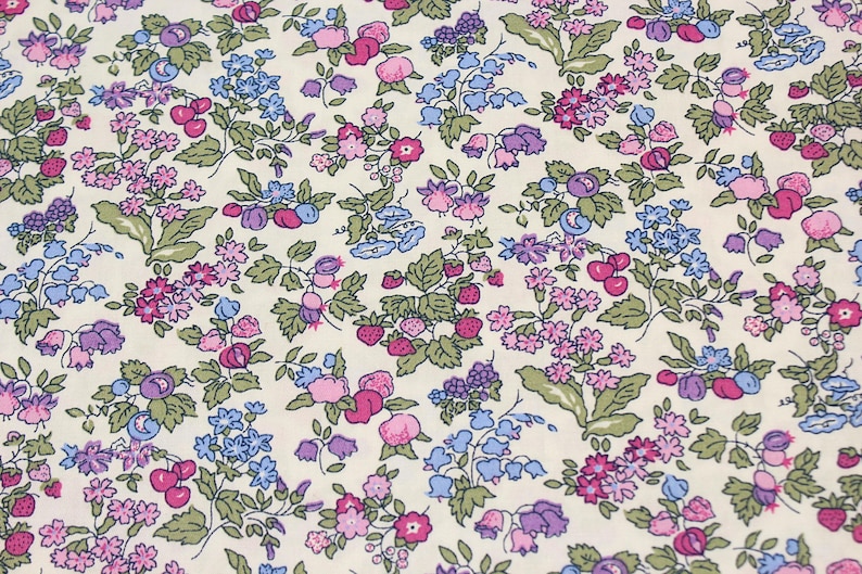 May include: A cream-colored fabric featuring a dense, repeating botanical print. The pattern consists of small pink, purple, and blue flowers, green leaves, and clusters of red strawberries and dark blue berries scattered across the surface.