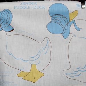 May include: A white fabric pattern for sewing a Jemima Puddle-Duck doll. The pattern includes a blue bonnet and a yellow beak. The text "Jemima Puddle-Duck" and "The World of Beatrix Potter" are printed on the fabric.
