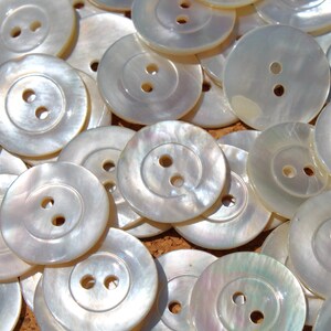 May include: A collection of round, pearlescent buttons with two holes each. The buttons have a subtle iridescent sheen, reflecting light. These sewing notions are ideal for various craft and apparel projects.