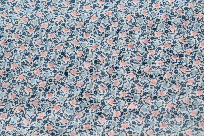 May include: Close-up of a fabric with a repeating floral pattern. The design features small pink and light blue flowers, along with dark blue stems and leaves, set against a white background. The fabric has a textured appearance.