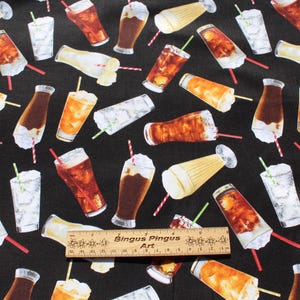 May include: Black fabric featuring a repeating pattern of drinks, including cola, iced tea, and milkshakes. The drinks are in glasses and bottles, with straws and whipped cream. A wooden ruler with text is at the bottom.