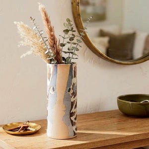 Könnte beinhalten: Eine zylindrische Keramikvase mit einem vertikalen beigen Mittelstreifen, der von blauen und braunen abstrakten Mustern flankiert wird. Die Vase ist mit getrockneten Pampasgraswedeln und grünen Eukalyptuszweigen gefüllt, was ein hohes Arrangement ergibt.