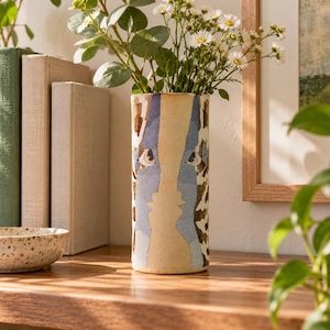 May include: A cylindrical ceramic vase featuring a cream-colored center panel with a blue silhouette of two faces looking at each other, flanked by brown and white giraffe-like patterns. The vase holds a bouquet of small white flowers with green stems.
