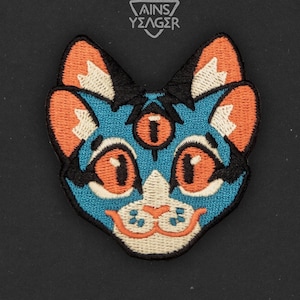 May include: Embroidered patch of a blue cat with orange ears, a white face, and three orange eyes. The cat has a small, white, smiling mouth.