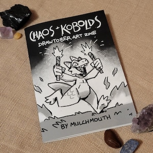 May include: A black and white zine titled "Chaos + Kobolds Drawtober Art Zine" featuring a cartoon dragon holding two lit sparklers. The zine cover has the text "By Mulchmouth" at the bottom. The zine is on a tan surface with crystals.