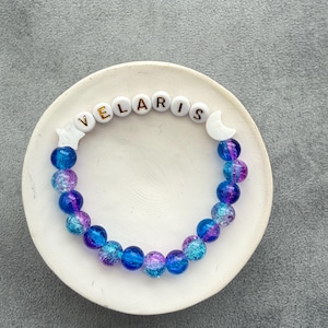 May include: A beaded bracelet featuring round, crackled glass beads in shades of blue and purple. The bracelet includes white letter beads spelling VELARIS, flanked by a white star-shaped bead on one side and a white crescent moon bead on the other.