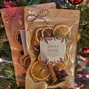 May include: Two clear pouches filled with dried orange slices, cinnamon sticks, and spices. Each pouch features a red and white twine bow and a label reading Holiday SIMMERING POTPOURRI with instructions to add ingredients to a saucepan.