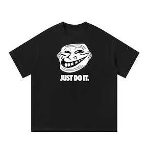 May include: A black short-sleeved t-shirt featuring a white graphic of the Trollface meme character centered on the chest. Below the graphic, the text "JUST DO IT." is printed in a bold, white, sans-serif font, mimicking a well-known brand slogan.