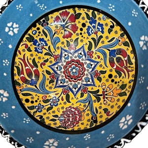 May include: A round ceramic bowl featuring a vibrant yellow center decorated with a multi-colored floral pattern and a central star motif. The wide light blue rim is adorned with small white flower accents and a black and white wavy border edge.