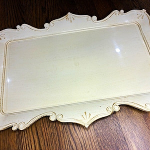 May include: A rectangular off-white serving tray with a raised, ornate, scalloped edge. The tray features a smooth, flat center surface and decorative, distressed gold-toned detailing along the intricate, curved border and corners.
