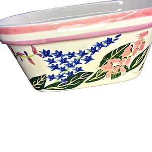 May include: An oval ceramic planter with a cream-colored base and a pink-painted rim. The exterior features a hand-painted botanical design with clusters of small blue star-shaped flowers, green leaves, and drooping pink bell-shaped blossoms.