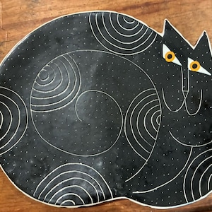 May include: A black ceramic plate shaped like a curled cat. The surface features white etched spiral patterns and small white dots. The cat has two pointed ears and two bright orange eyes with white triangular accents, outlined in thin white lines.