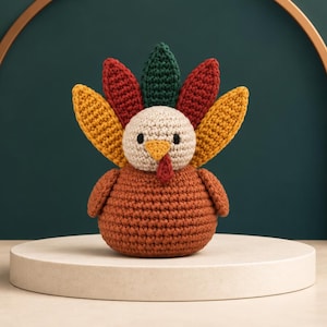 Op de afbeelding: Een gehaakt kalkoenfiguurtje met een ronde beige kop, een bruin lichaam en een waaier van kleurrijke staartveren in de tinten geel, rood en donkergroen. De kalkoen heeft een kleine oranje snavel, een rode lel en twee kleine zwarte ogen.